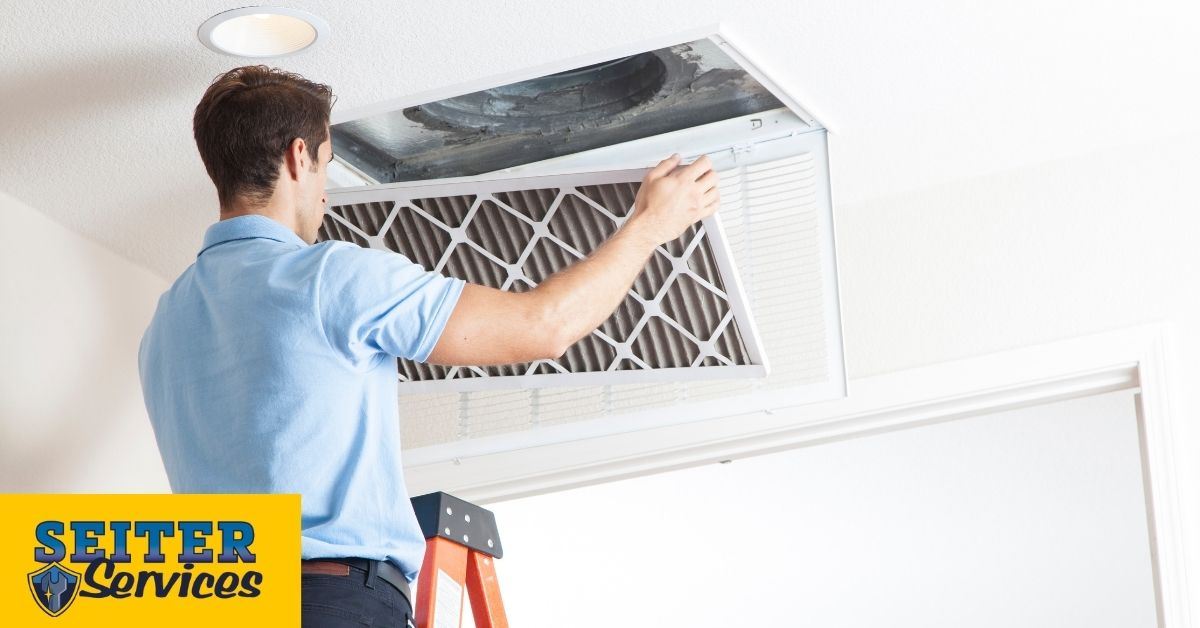 A man holding an air filter below a ceiling air vent with the vent door open